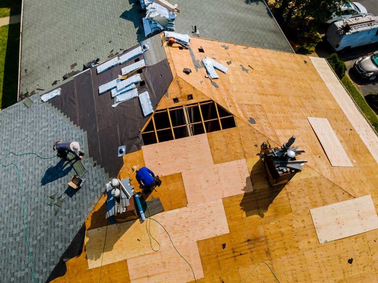 Two workers installing composite shingles on a house roof under partly cloudy skies.
