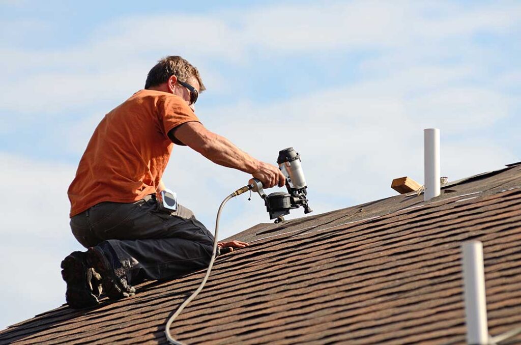 A homeowner observes a roofer inspecting shingles and flashing on a two-story house during clear fall weather.