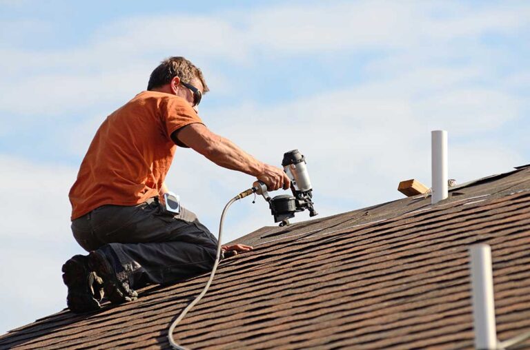A homeowner observes a roofer inspecting shingles and flashing on a two-story house during clear fall weather.